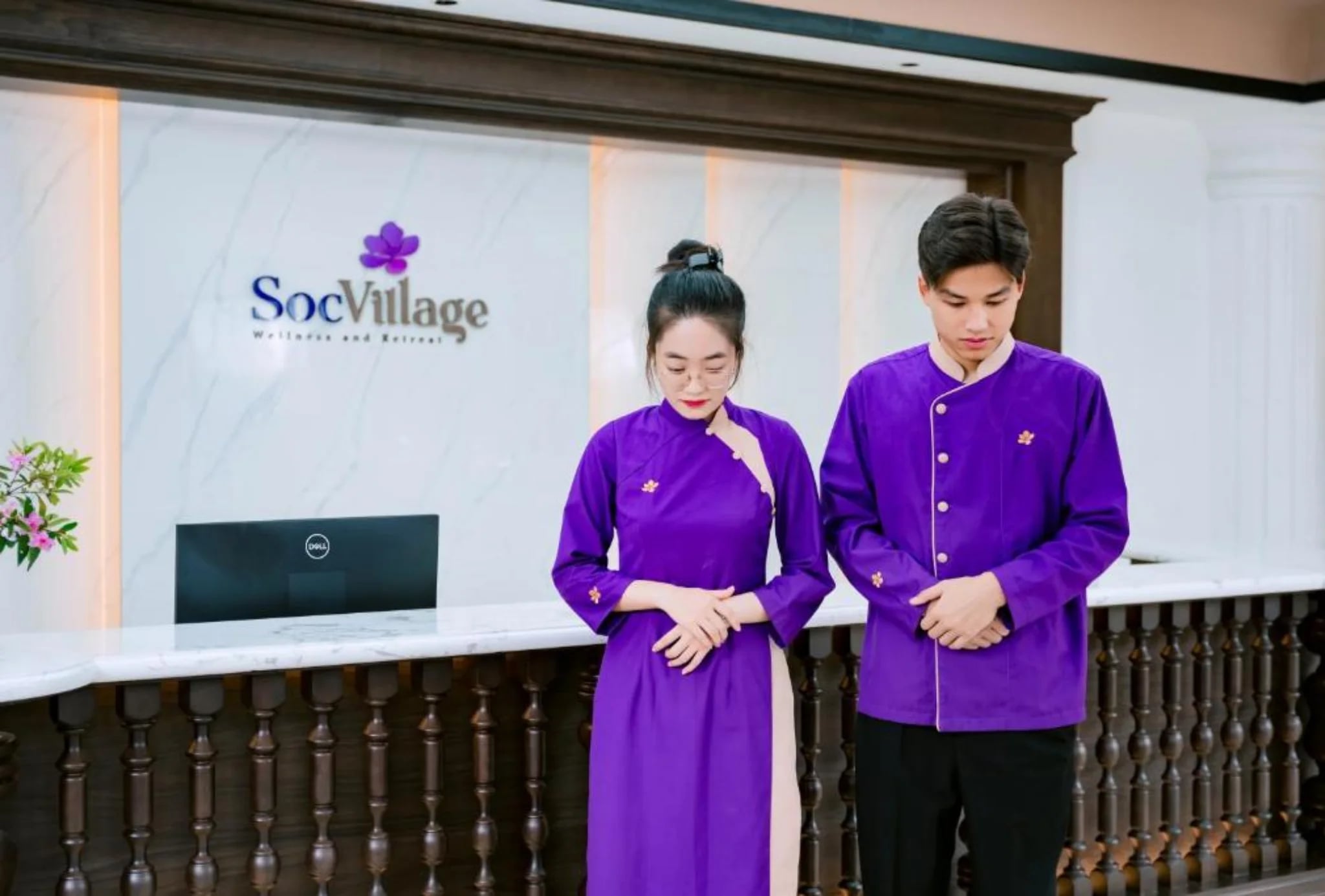 Hai nhân viên lễ tân mặc đồng phục tím cúi chào tại quầy Soc Village Wellness & Retreat